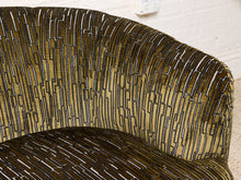 Load image into Gallery viewer, Image 11 of 16: Close-up of a curved upholstered armchair with a textured patterned fabric in olive green, black, and white, set against a white brick wall background.

