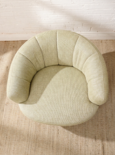 Load image into Gallery viewer, Image 6 of 16: A round, low-profile upholstered accent chair in a textured light sage green fabric, set on a woven jute rug against a white brick wall.
