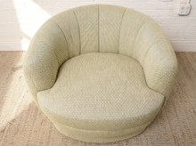 Load image into Gallery viewer, Image 7 of 16: A round, low-profile barrel chair upholstered in a textured light sage green fabric, set on a woven jute rug against a white brick wall.
