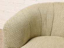 Load image into Gallery viewer, Image 8 of 16: Close-up of a curved upholstered armchair in a light beige fabric with a small repeating geometric pattern, set against a white brick wall background.
