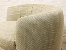 Load image into Gallery viewer, Image 11 of 16: Close-up of a curved upholstered armchair in a light beige fabric with a small repeating geometric pattern, set against a white brick wall.
