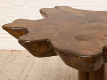 Load image into Gallery viewer, Image 6 of 16: A close-up of a rustic, hand-carved wooden stool with a natural, irregular leaf-like silhouette and a glossy dark brown finish, set against a white brick wall and light carpet.
