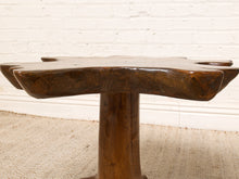 Load image into Gallery viewer, Image 11 of 16: A rustic, hand-carved wooden table with a thick, irregularly shaped top and a single central pedestal base, shown against a white brick wall and light-colored rug.
