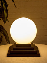 Load image into Gallery viewer, Image 2 of 22: A glowing spherical table lamp with a warm white frosted globe and a dark wood tiered base, set on a textured beige surface against a warm brick wall with a plant leaf visible on the left.
