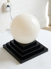 Load image into Gallery viewer, Image 3 of 22: A modern table lamp featuring a large, glossy white spherical globe on a tiered black wooden base, set on a white textured surface against a white brick wall.
