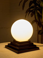 Load image into Gallery viewer, Image 6 of 22: A modern table lamp with a glowing white spherical shade on a tiered dark brown wooden base, set on a textured beige surface with a warm, dimly lit background and a plant silhouette.

