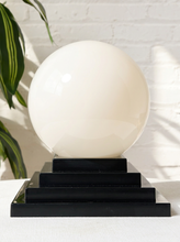 Load image into Gallery viewer, Image 7 of 22: A large glossy white spherical crystal ball displayed on a tiered black pedestal base, set against a white brick wall with soft leaf shadows and a green plant visible on the left.
