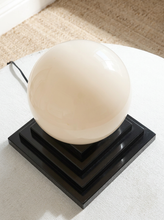 Load image into Gallery viewer, Image 9 of 22: A modern table lamp featuring a large, glossy cream-colored spherical shade resting on a tiered black square base, set on a white textured surface.

