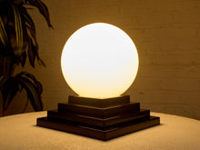Load image into Gallery viewer, Image 12 of 22: A glowing spherical table lamp with a warm white frosted globe resting on a dark tiered wooden base, set on a textured beige surface against a softly lit brick wall with a plant leaf visible on the left.
