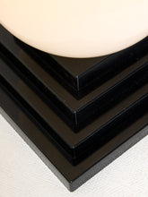 Load image into Gallery viewer, Image 15 of 22: Close-up of a glossy black tiered pedestal base with a smooth off-white rounded top surface, photographed on a light textured fabric background.
