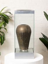 Load image into Gallery viewer, Image 1 of 18: Vintage dark brown sculptural vase with rounded shoulders, a narrow circular opening, tapered body, and weathered surface, displayed inside a tall rectangular glass case with brushed silver-tone metal top and base.

