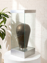 Load image into Gallery viewer, Image 2 of 18: Vintage dark brown sculptural vase with rounded shoulders, a narrow circular opening, tapered body, and weathered surface, displayed inside a tall rectangular glass case with brushed silver-tone metal top and base.
