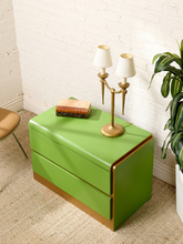 Load image into Gallery viewer, Image 3 of 16: A bright green two-drawer nightstand with gold trim, styled in a room corner with a brass two-light candelabra, books, a potted plant, and a white brick wall background.
