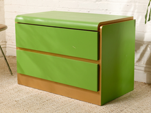 Load image into Gallery viewer, Image 5 of 16: A mid-century modern two-drawer nightstand with a glossy lime green finish and a warm tan wood base, shown in a room setting with a white brick wall and carpet.

