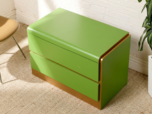 Load image into Gallery viewer, Image 6 of 16: A bright green two-drawer nightstand with a glossy finish and a metallic gold base trim, shown in a styled room setting with a white brick wall, beige carpet, and a plant.
