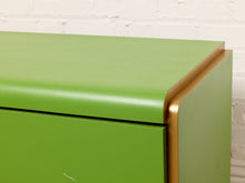 Load image into Gallery viewer, Image 9 of 16: Close-up of a bright green cabinet or dresser showing the top surface, front drawer, and side panel with a metallic gold trim, set against a white brick wall.
