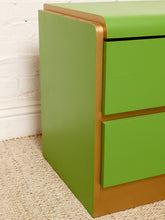 Load image into Gallery viewer, Image 10 of 16: A close-up of a modern two-drawer nightstand or cabinet with a bright lime green finish and a metallic gold-toned frame, shown against a white brick wall and beige carpet.
