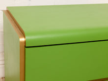 Load image into Gallery viewer, Image 11 of 16: Close-up of a bright green cabinet or dresser drawer with a curved front edge and metallic gold trim, set against a white brick wall background.
