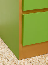 Load image into Gallery viewer, Image 12 of 16: Close-up of a green drawer unit corner with a warm tan wood baseboard, shown on a woven beige rug.
