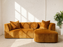 Load image into Gallery viewer, Image 1 of 16: Bonnie Two-Piece Sectional Sofa in Thriller Amber featuring golden amber textured upholstery, rounded arms, generously cushioned seating, a right-facing curved chaise, two square accent pillows, and a matching round tufted pillow.
