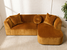 Load image into Gallery viewer, Image 2 of 16: Bonnie Two-Piece Sectional Sofa in Thriller Amber featuring golden amber textured upholstery, rounded arms, generously cushioned seating, a right-facing curved chaise, two square accent pillows, and a matching round tufted pillow.
