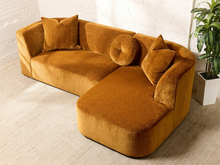 Load image into Gallery viewer, Image 4 of 16: Bonnie Two-Piece Sectional Sofa in Thriller Amber featuring golden amber textured upholstery, rounded arms, generously cushioned seating, a right-facing curved chaise, two square accent pillows, and a matching round tufted pillow.
