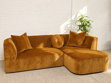 Load image into Gallery viewer, Image 5 of 16: Bonnie Two-Piece Sectional Sofa in Thriller Amber featuring golden amber textured upholstery, rounded arms, generously cushioned seating, a right-facing curved chaise, two square accent pillows, and a matching round tufted pillow.
