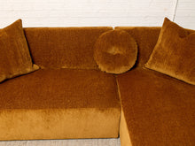Load image into Gallery viewer, Image 10 of 16: A mustard yellow velvet sectional sofa with two matching square throw pillows and a round round throw pillow, set against a white brick wall.

