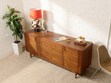 Load image into Gallery viewer, Image 4 of 24: Vintage mid-century modern walnut credenza by Kroehler Furniture featuring three central drawers with brass-tone horizontal pulls, two side cabinet doors with vertical handles, richly figured walnut wood grain, and slender tapered wooden legs.

