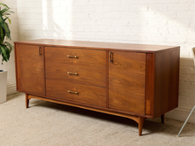 Load image into Gallery viewer, Image 1 of 24: Mid-century modern wooden sideboard cabinet with three drawers and two doors, featuring brass handles, set against a white brick wall and beige carpet.
