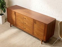 Load image into Gallery viewer, Image 6 of 24: Mid-century modern wooden sideboard cabinet with three drawers and two doors, shown in a styled room corner with a white brick wall, beige carpet, and a potted plant.
