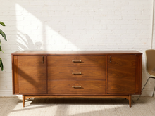 Load image into Gallery viewer, Image 5 of 24: Vintage mid-century modern walnut credenza by Kroehler Furniture featuring three central drawers with brass-tone horizontal pulls, two side cabinet doors with vertical handles, richly figured walnut wood grain, and slender tapered wooden legs.
