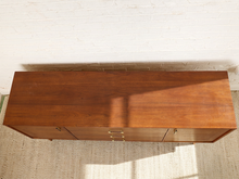 Load image into Gallery viewer, Image 7 of 24: A mid-century modern wooden sideboard or credenza with a warm medium-brown wood finish and brass drawer pulls, photographed from a high angle against a white brick wall and textured rug.
