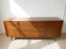 Load image into Gallery viewer, Image 8 of 24: Mid-century modern wooden sideboard with three drawers and two cabinet doors, shown against a white brick wall on a light rug.
