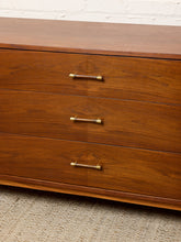 Load image into Gallery viewer, Image 11 of 24: A close-up of a mid-century modern wooden dresser with three drawers, featuring warm brown wood grain and brass cylindrical handles, set against a white brick wall and textured carpet.
