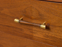 Load image into Gallery viewer, Image 13 of 24: A close-up of a mid-century modern wooden drawer pull handle with a dark wood center and brass-toned ends, mounted on a warm brown wood cabinet door.
