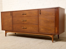 Load image into Gallery viewer, Image 17 of 24: Mid-century modern wooden sideboard credenza with three drawers and two cabinet doors, featuring brass handles and tapered legs, set against a white brick wall and beige carpet.
