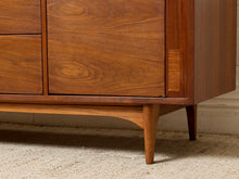 Load image into Gallery viewer, Image 18 of 24: Close-up of a mid-century modern wooden sideboard cabinet with warm brown walnut grain, tapered legs, and a slatted wood drawer front, shown on a beige carpet.
