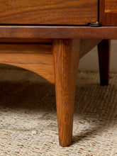 Load image into Gallery viewer, Image 19 of 24: Close-up of a mid-century modern wooden side table or cabinet leg with a tapered wood finish, resting on a textured beige woven rug.
