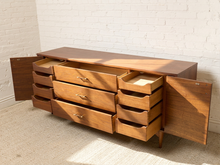 Load image into Gallery viewer, Image 21 of 24: Mid-century modern wooden credenza with multiple drawers and open cabinet doors, set against a white brick wall and light-colored carpet.
