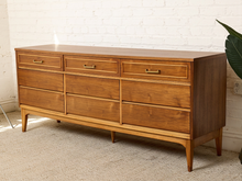Load image into Gallery viewer, Image 5 of 21: Mid-century modern wooden dresser with six drawers, brass handles, and tapered legs, positioned against a white brick wall on a beige carpet.
