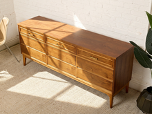 Load image into Gallery viewer, Image 6 of 21: A mid-century modern wooden dresser with six drawers and brass handles, positioned against a white brick wall with warm sunlight casting shadows on the floor.
