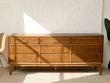 Load image into Gallery viewer, Image 4 of 21: A mid-century modern wooden dresser with six drawers and brass handles, positioned against a white brick wall with diagonal sunlight shadows.
