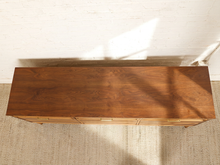 Load image into Gallery viewer, Image 7 of 21: A mid-century modern wooden sideboard or credenza with a warm walnut finish, featuring multiple drawers with brass pulls, photographed from a top-down angle against a white brick wall and textured rug.
