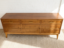 Load image into Gallery viewer, Image 8 of 21: A mid-century modern wooden sideboard with six drawers, featuring a warm medium-brown wood finish and brass pull handles, set against a white brick wall and light beige rug.
