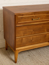 Load image into Gallery viewer, Image 9 of 21: Mid-century modern wooden chest of drawers with a warm medium-brown finish, featuring a single top drawer with a brass pull handle and two larger drawers below, set against a white brick wall and textured beige carpet.
