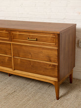 Load image into Gallery viewer, Image 11 of 21: Mid-century modern wooden dresser with a warm medium-brown finish, brass hardware, and tapered legs, shown in a room setting against a white brick wall and textured carpet.
