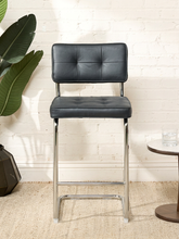 Load image into Gallery viewer, Image 3 of 17: 1970s-style counter stool featuring black button-tufted upholstered seat and backrest, polished chrome tubular cantilever frame, curved sled-style base, and integrated metal footrest.
