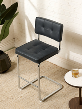 Load image into Gallery viewer, Image 2 of 17: 1970s-style counter stool featuring black button-tufted upholstered seat and backrest, polished chrome tubular cantilever frame, curved sled-style base, and integrated metal footrest.
