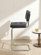 Load image into Gallery viewer, Image 4 of 17: 1970s-style counter stool featuring black button-tufted upholstered seat and backrest, polished chrome tubular cantilever frame, curved sled-style base, and integrated metal footrest.
